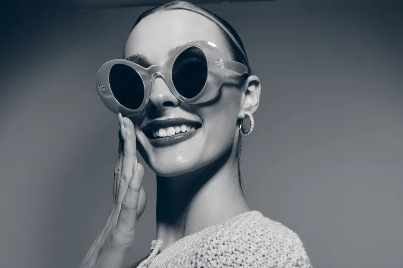Smiling young Caucasian woman with pale skin wearing large round sunglasses and knitted sweater touches her cheek in a studio with soft lighting. Concept for fashion and style.