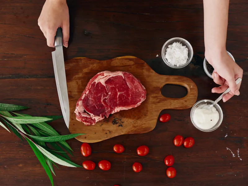 Женщина с ножом в руке нарезает свежее стейковое мясо для жарки на кухне
