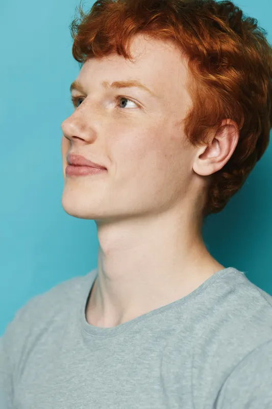 Young man with red hair and fair skin looking sideways wearing gray shirt. Portrait with soft natural light on blue background for casual lifestyle and positive expression.