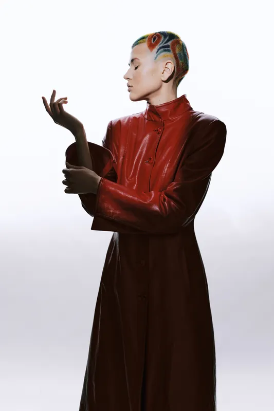 Creative woman with colorful shaved hair wearing a long red leather coat, posing elegantly against a bright white background in a modern studio setting.