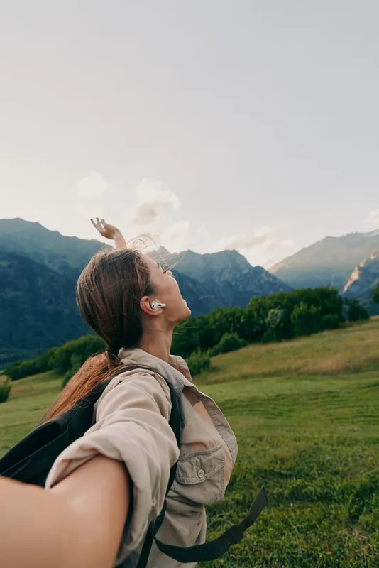 Cheerful woman outdoors, arms raised, enjoying mountain scenery, backpack ready, sunlit moment, fresh breeze, natural colors, expansive valley background