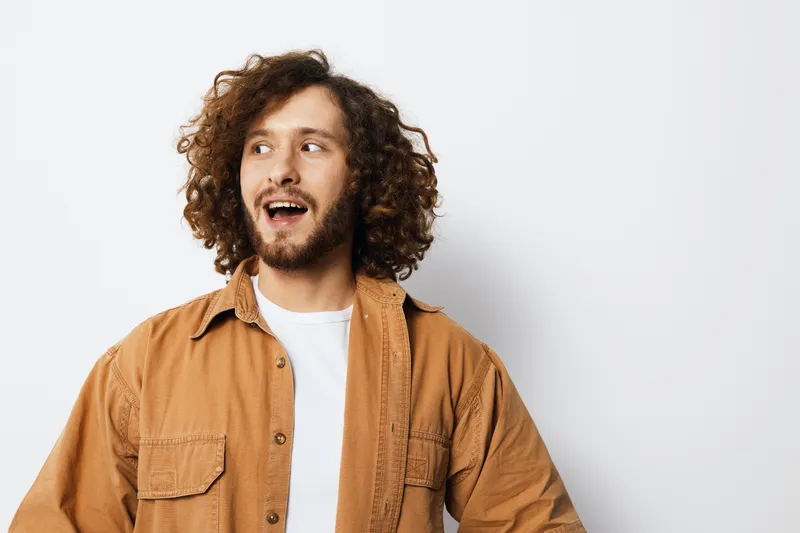 Cheerful young man with curly hair wearing a casual outfit, expressing joy against a light background, showcasing a trendy seasonal style