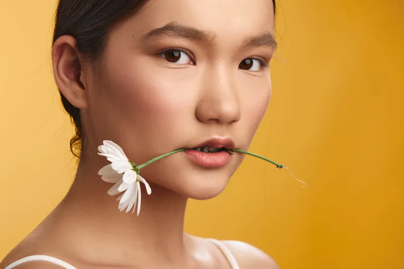 asian woman with a flower in her mouth stands against a warm yellow backdrop, exuding calm elegance and poise, a simple yet striking portrait of natural beauty.
