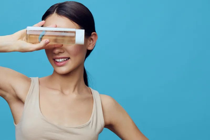 A cheerful young woman of Asian descent playfully holds a transparent container while posing against a blue background, showcasing a vibrant and fresh concept