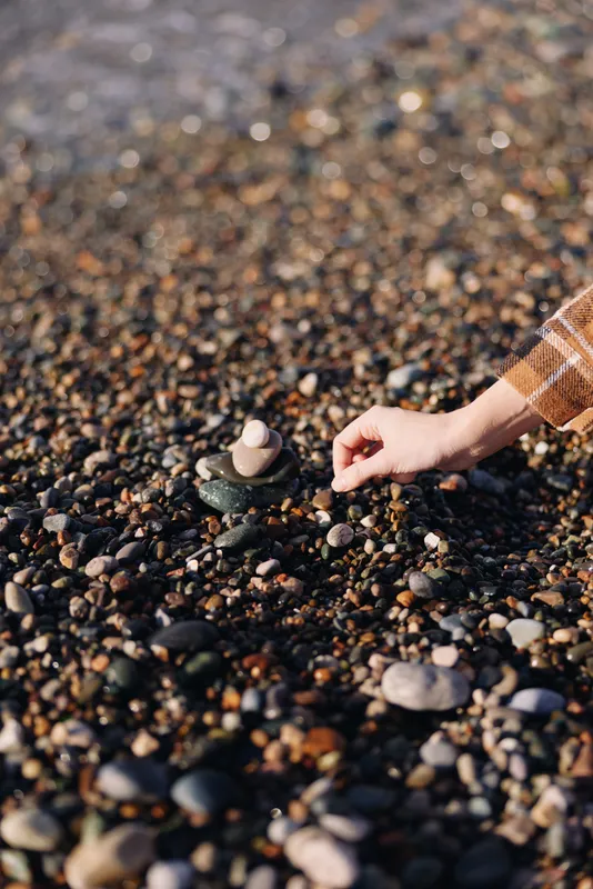 Hand placing a small stone on a pebble beach, creating a delicate stack among smooth rocks and coarse sand, capturing calm, natural balance and tranquil outdoor scenery at golden hour
