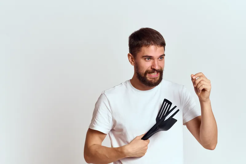 Мужчина с кухонными приборами в руках, в белой футболке, на светлом фоне, обрезанный вид