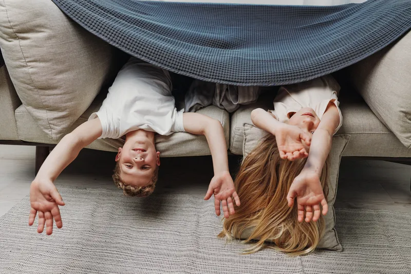 Two happy children lying upside down on a couch under a blanket, smiling and reaching out with their hands. Cozy home environment with playful and joyful mood.