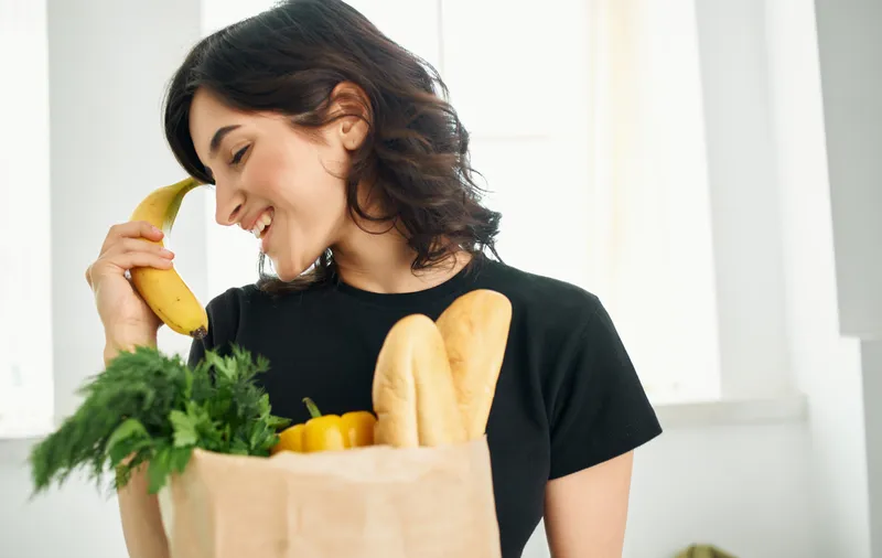 Брюнетка в чёрной футболке несёт пакет с продуктами, покупая здоровую пищу