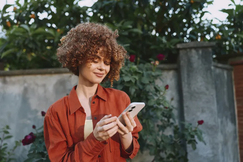 A woman with curly hair in an orange shirt stands outdoors, gazing at a smartphone with a thoughtful smile. A casual lifestyle moment captured in bright natural light for everyday connectivity