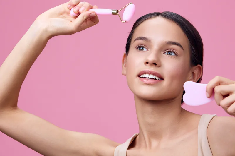 Young woman smiling while using skincare tools, showcasing healthy skincare routine and natural beauty against a pink background