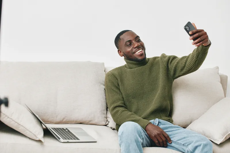 Smiling African man taking a selfie on the couch with a laptop nearby, wearing a green sweater, relaxing at home, joyful mood and casual lifestyle