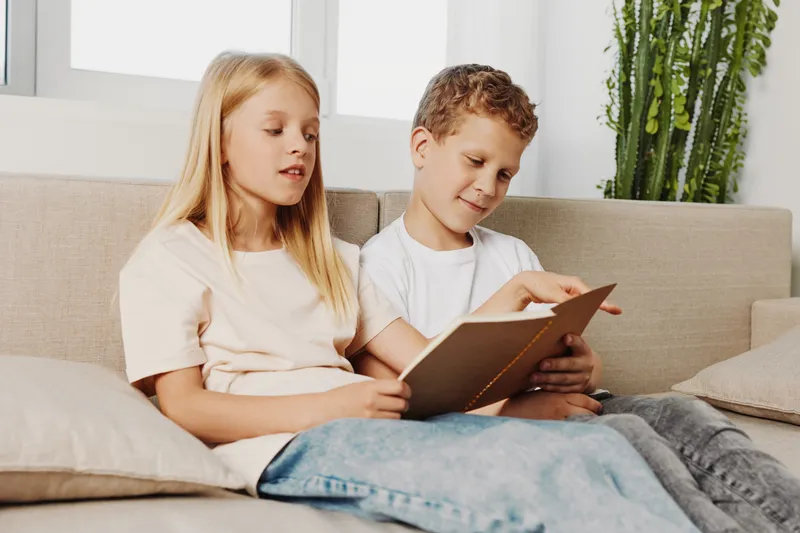 Two children sit on a sofa reading a book together in a bright room with plants. Concept of childhood, education, learning, and family bonding moments.