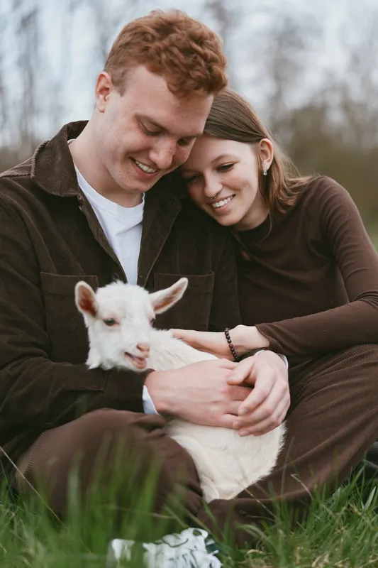 Young couple sitting on grass gently holding a white baby goat while sharing a tender moment outdoors in casual brown clothing during a calm day.