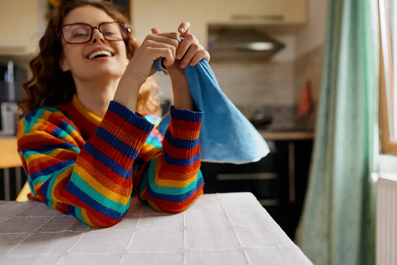 Young woman with curly hair cleans a blue cloth on a kitchen table, smiling while tidying and enjoying a cozy home moment.
