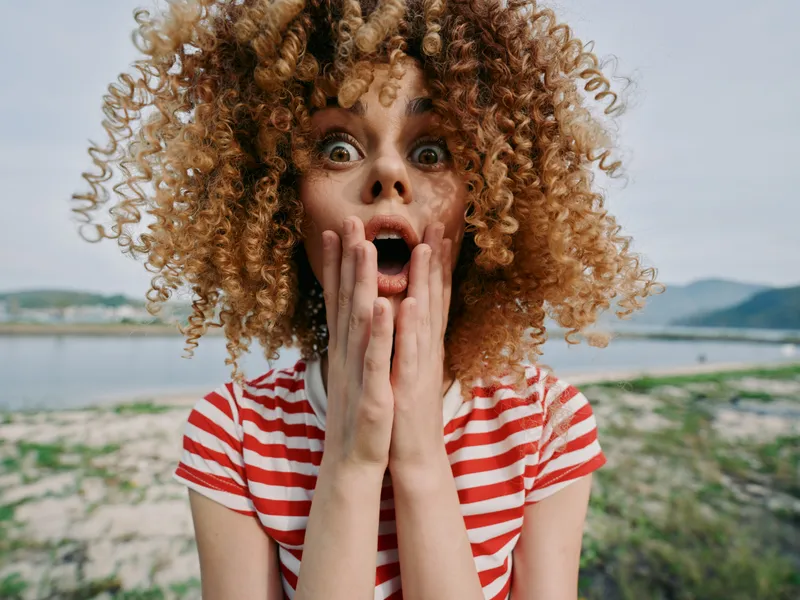 Young woman with voluminous curly hair and wide eyes presses hands to face in astonishment on a bright beach scene, striped shirt, natural light, candid emotion.