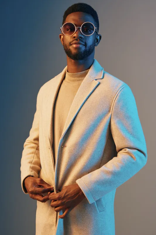 Stylish Black male model in beige coat wearing round glasses against a blue and gray background, exuding confidence and modern fashion, perfect for fashion and lifestyle themes