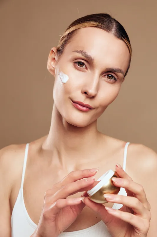 Young woman with blonde hair applies cream to her cheek in soft studio lighting, a calm skincare moment, relaxed posture, and a clean neutral background