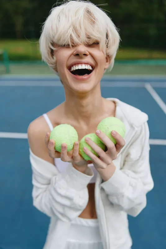 Happy female tennis player holding green tennis balls on a blue court, wearing casual sporty attire, expressing joy and enthusiasm in a sunny outdoor setting