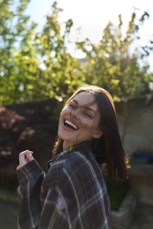 Cheerful woman outdoors, smiling with plaid shirt, sunlit moment, relaxed posture, natural colors, leafy background, candid portrait, joyful atmosphere