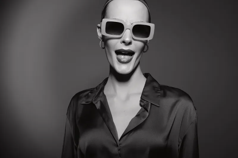 Stylish white woman with pale skin wearing large sunglasses and dark blouse poses confidently in monochrome studio portrait.