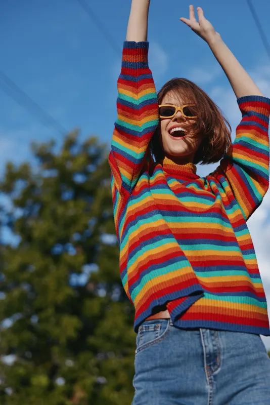 Woman in rainbow sweater and sunglasses raises her arms outdoors smiling happily, portrait of joy and casual fashion, vibrant striped knit with blue sky background and carefree energy