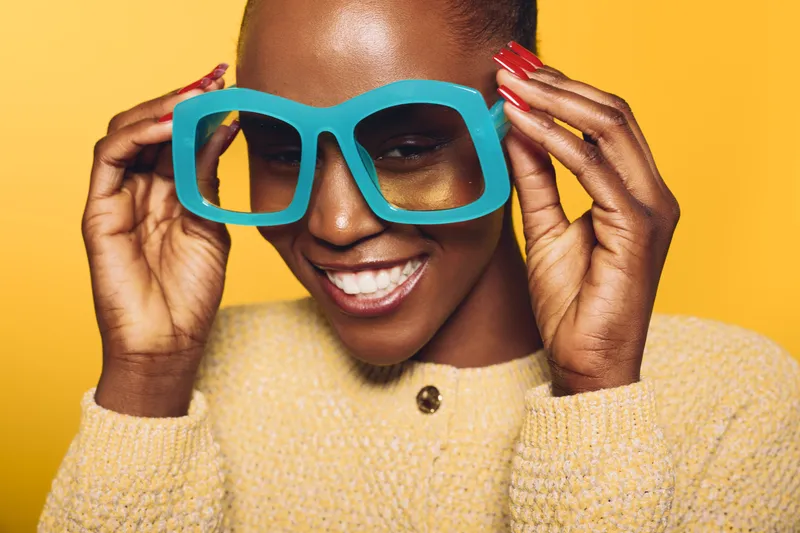 Happy dark skinned African woman in yellow sweater holds oversized blue glasses on yellow background. Close-up portrait with bright colors and cheerful expression for fashion concept.
