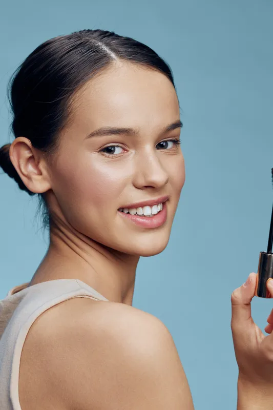 Young woman with clear skin holds mascara smiling against a light blue background, embodying beauty and confidence in cosmetics