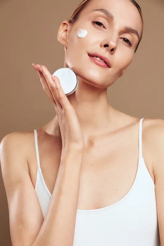 Young woman with clean skin holds open jar of face cream near cheek with small amount of product applied, wearing white tank top, isolated on beige background with soft lighting.