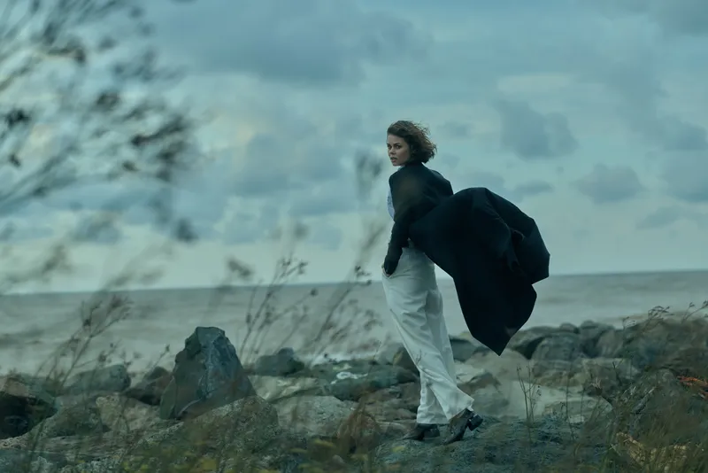 windy coastline scene with a woman walking along rocky shore, flowing coat catching the breeze, ocean horizon in the distance, moody blue tones and natural outdoor atmosphere