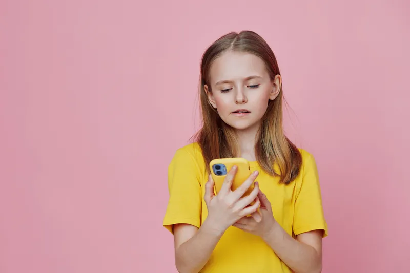 A young girl with long hair wearing a bright yellow t shirt is focused on her smartphone, showcasing emotions of concentration against a soft pink background