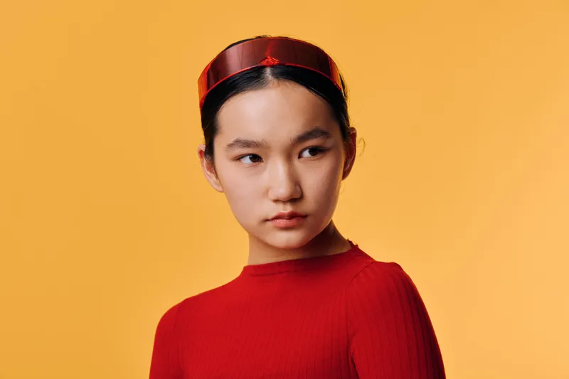 Young woman in red sweater and matching headband looks sideways against orange background with serious expression for fashion and style portrait closeup.