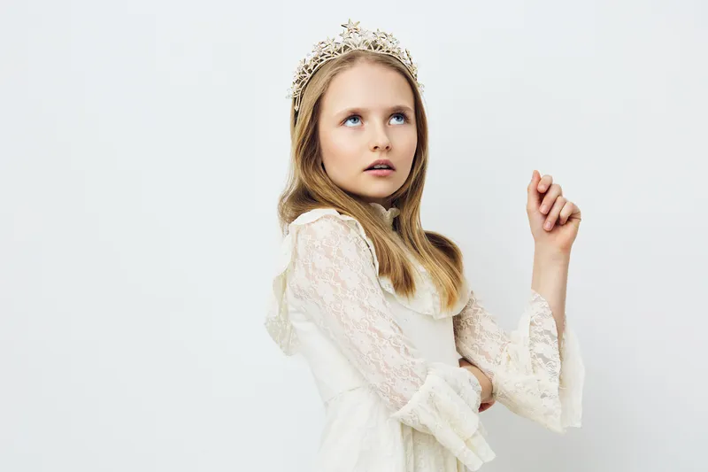 Elegant young girl adorned with a crown, showcasing innocence and wonder against a soft, light background Styled in white lace dress, conveying a sense of beauty and imagination