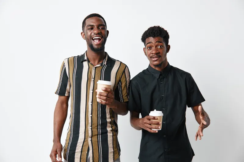 Two young black men with dark skin holding coffee cups smiling and standing together on white background in casual and formal shirts.