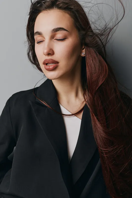 Young woman with long hair and closed eyes wearing black jacket and white shirt, posing indoors against grey wall with natural makeup and relaxed expression.