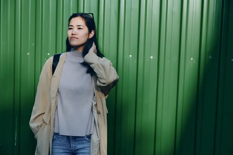 Woman in a beige coat and gray sweater stands against a green corrugated wall, casual fashion scene, thoughtful pose, urban backdrop, natural light, everyday style, lifestyle photography for