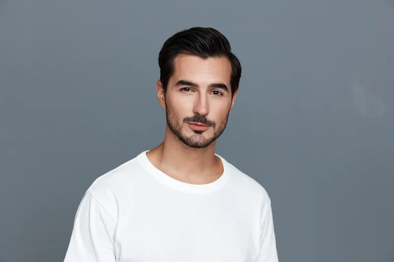 Handsome young man in a white t-shirt poses confidently against a gray background, showcasing a charming smile and modern hairstyle. Portrait concept for lifestyle.
