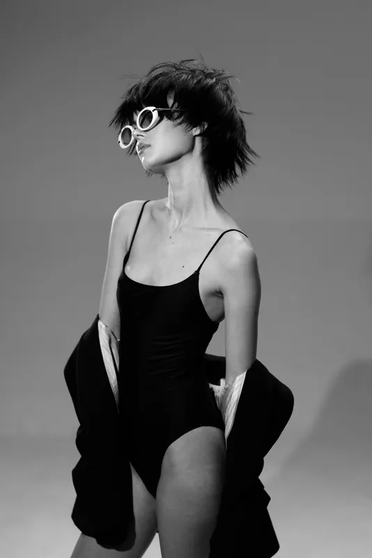 Stylish woman with sunglasses in black swimsuit and jacket posing in black and white studio portrait.