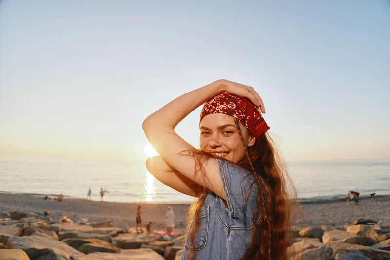 Lifestyle woman in modern boho-western denim street style smiling with red bandana by the beach at sunset, warm film color tones enhancing free-spirited vibe.