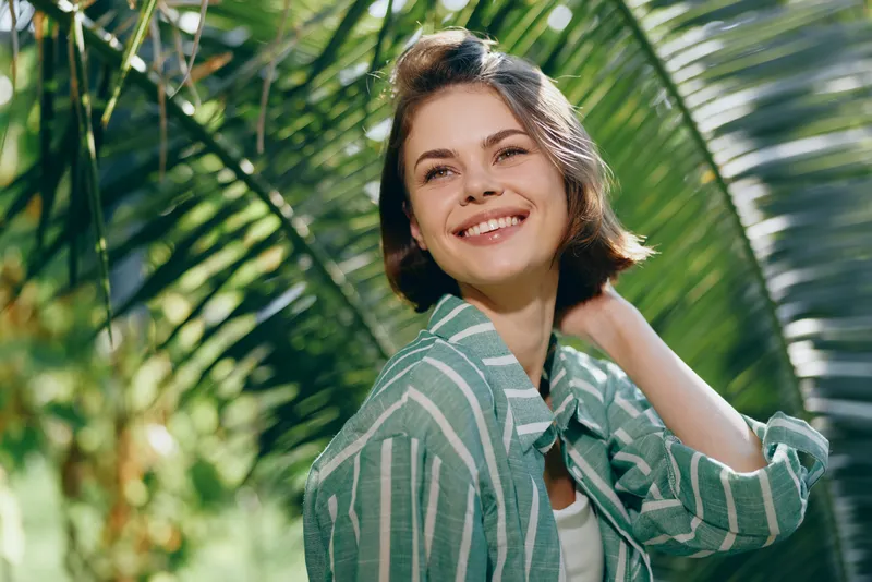 Bright portrait of a cheerful woman in a striped green shirt, standing among lush tropical light and vibrant foliage.