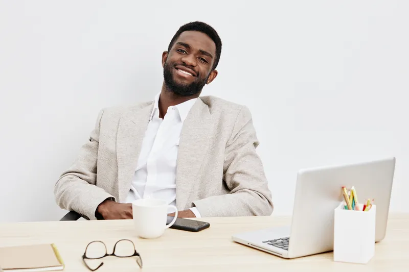 Smiling man, professional, business attire, seated at desk with laptop, coffee mug, glasses, modern office background. Casual confident portrait of a cheerful businessman working in a corporate enviro