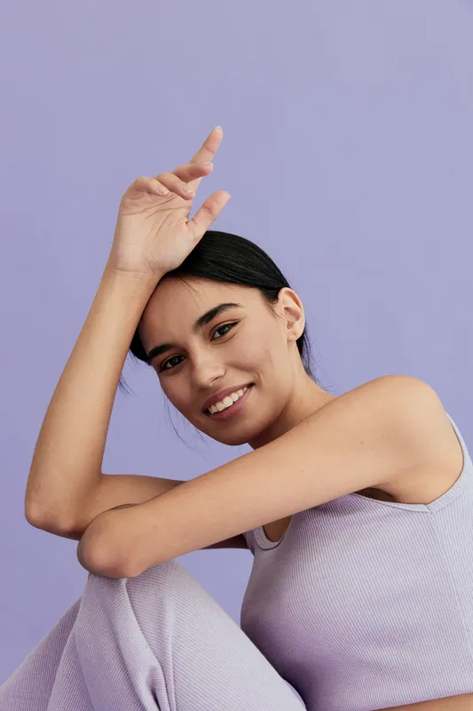smiling woman with dark hair wearing lilac sleeveless top and pants poses indoors against pastel purple background in casual and relaxed style.