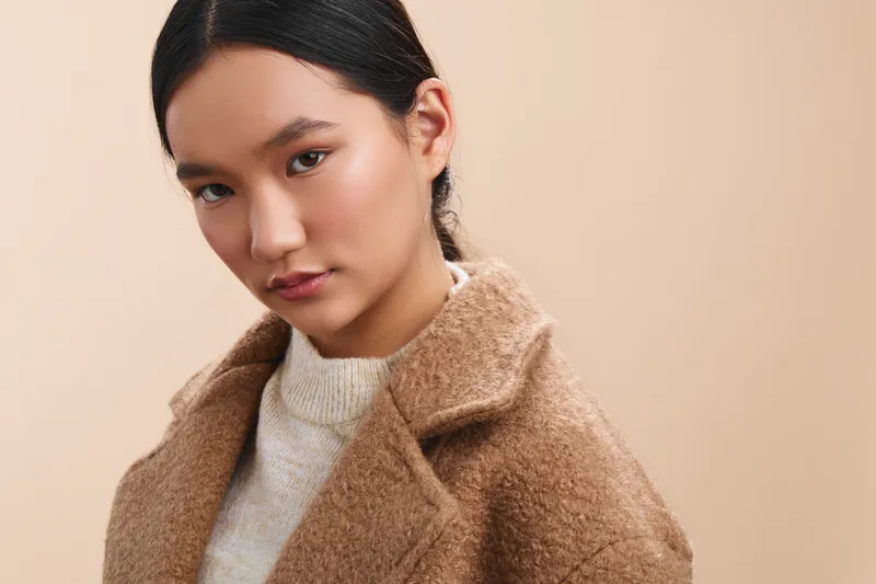 asian woman wears a tan wool coat and neutral sweater, gazing softly at the camera with a calm, warm presence, creating a minimalist fashion portrait with gentle tones and refined mood.