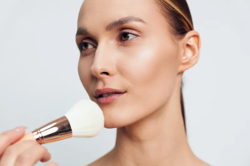 A young woman applies powder makeup on her face with a large brush, focusing on flawless skin and natural beauty. Concept for skincare, cosmetics, and daily makeup routine.