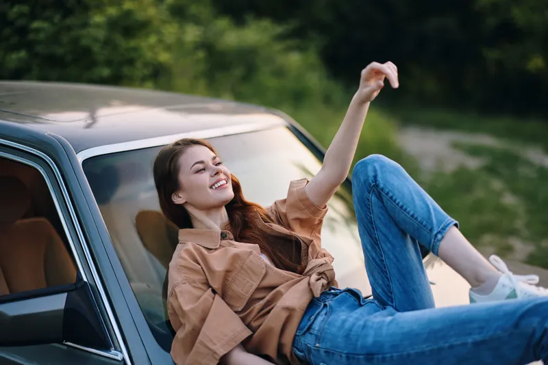 Young woman enjoying a sunny day, relaxing on a car hood in casual attire, showcasing a joyful expression in a green outdoor setting.