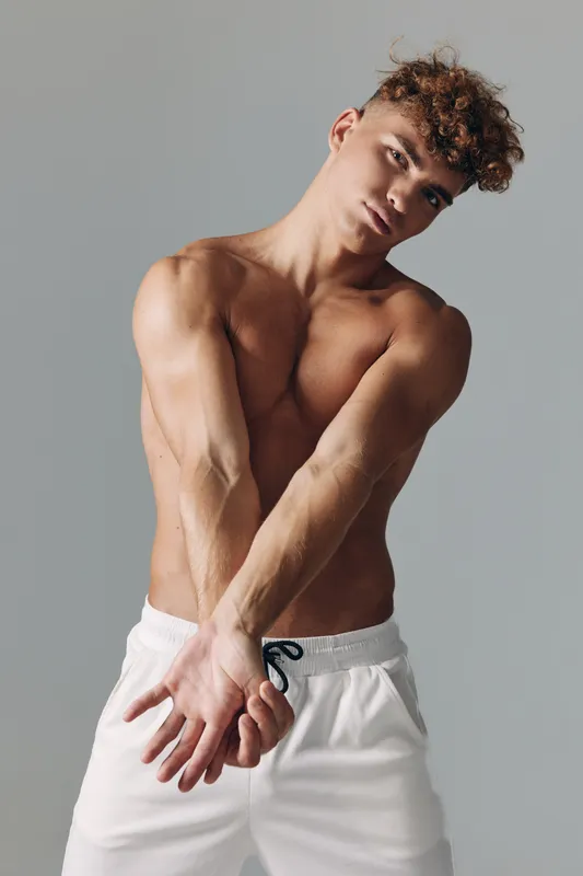 Young muscular man with curly hair stretching arms forward shirtless wearing white shorts against plain background for fitness and workout concept.