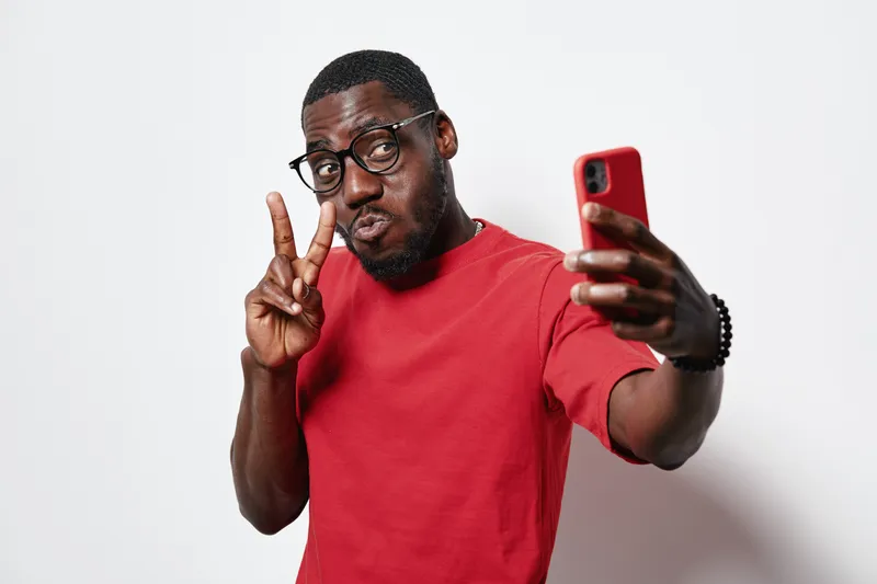 Black man with dark skin wearing glasses and red t shirt taking selfie with smartphone showing peace sign on white background. Casual style and confident expression in studio.