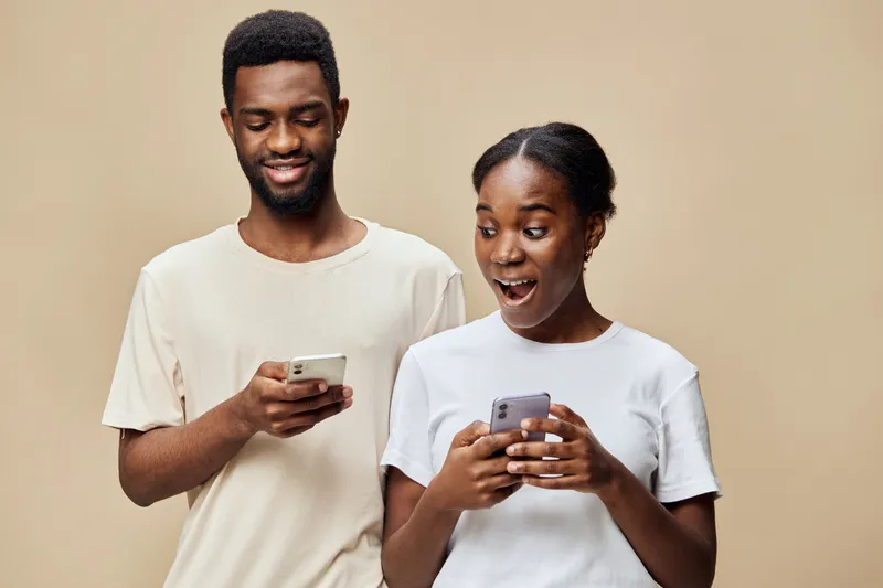 Happy young African couple sharing exciting news on smartphones, expressing joy and engagement in a modern lifestyle setting with neutral background