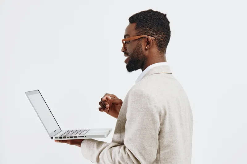 Professional man working on laptop device with digital technology, wearing light blazer and glasses, standing against plain white background, business and workspace concept.