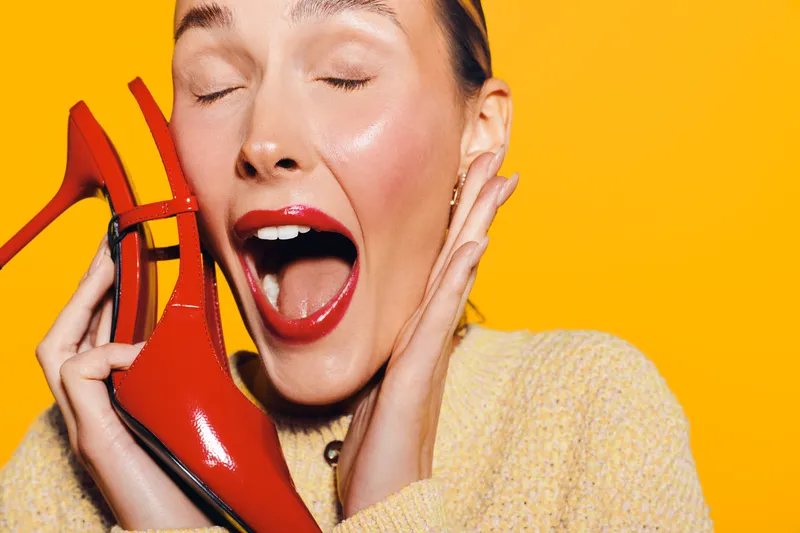 Happy woman with red lipstick holding red high heel shoe near face, eyes closed and smiling. Bright yellow background and cozy sweater create cheerful fashion concept.
