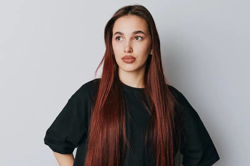 Young woman with long straight hair wearing black shirt standing with hands on hips looking away on light background. Casual style, confident pose, natural makeup and relaxed expression.